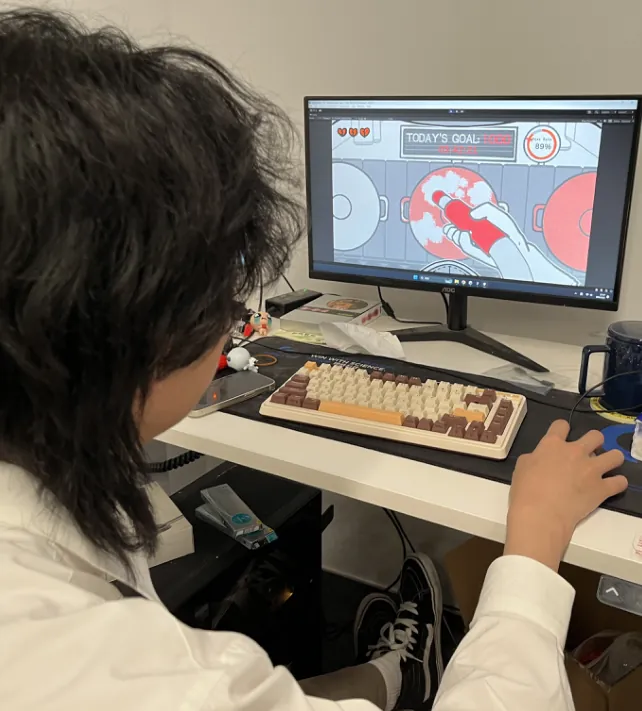 A player with curly hair sitting at a desk, playing Sanding Man on a computer monitor.