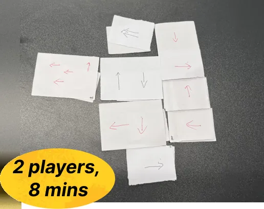 Paper prototype cards with hand-drawn red arrows spread on a dark table, with a yellow badge reading 2 players, 8 mins.