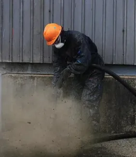 A worker in an orange hard hat and mask sandblasting beside a wall as clouds of dust rise.